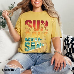 May include: Mustard yellow t-shirt with the words "SUN SALT SAND" in a retro design. The graphic features palm trees, waves, and sand. The shirt is a relaxed fit and has short sleeves.