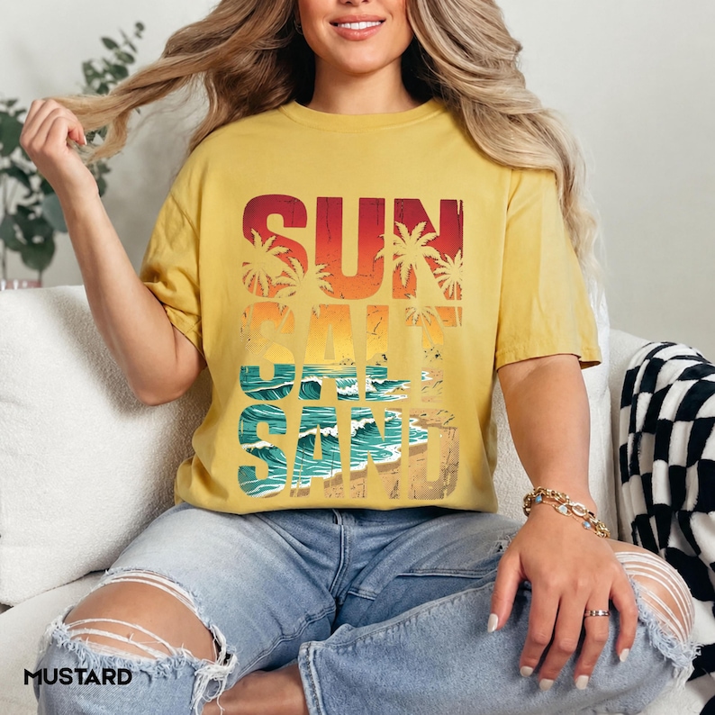 May include: Mustard yellow t-shirt with the words "SUN SALT SAND" in a retro design. The graphic features palm trees, waves, and sand. The shirt is a relaxed fit and has short sleeves.