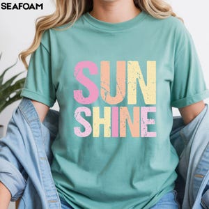 Seafoam green t-shirt with 'SUNSHINE' graphic in distressed lettering, 'SUN' in pink and orange, 'SHINE' in yellow and pink