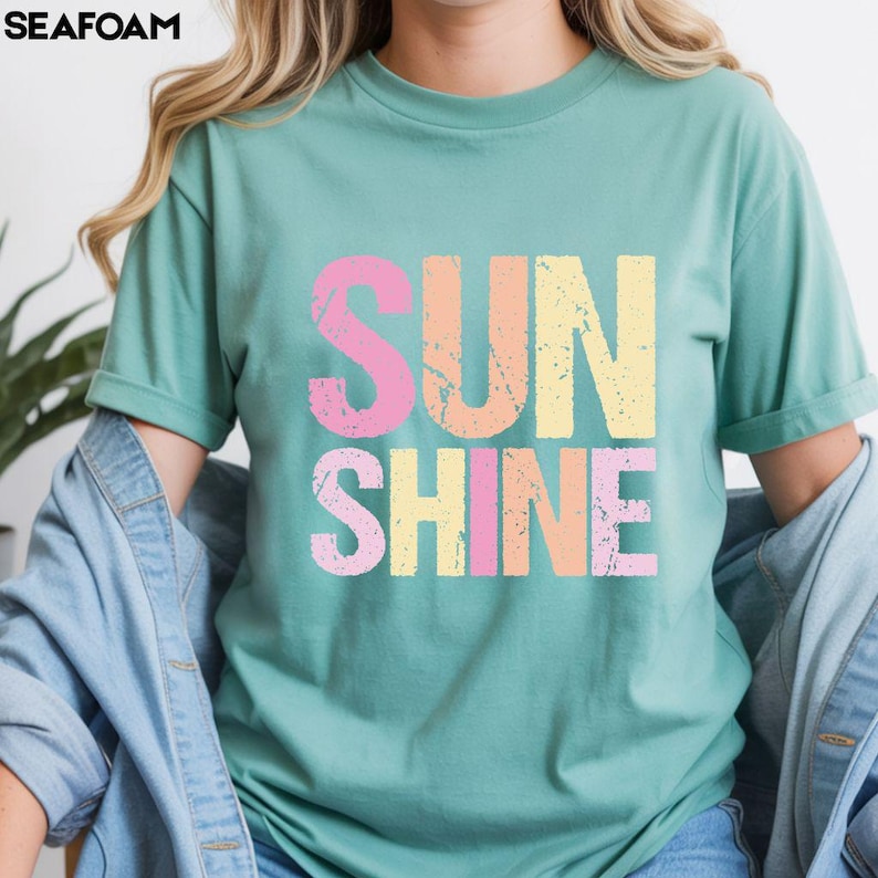 Seafoam green t-shirt with 'SUNSHINE' graphic in distressed lettering, 'SUN' in pink and orange, 'SHINE' in yellow and pink