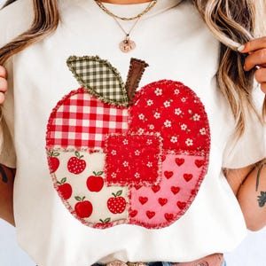 Może przedstawiać: Kremowy t-shirt z dużą, centralną aplikacją w kształcie jabłka wykonaną z różnych kawałków materiału. Wzór jabłka zawiera czerwono-białą kratkę, czerwone motywy kwiatowe, małe czerwone jabłka oraz czerwone serca z postrzępionymi brzegami.