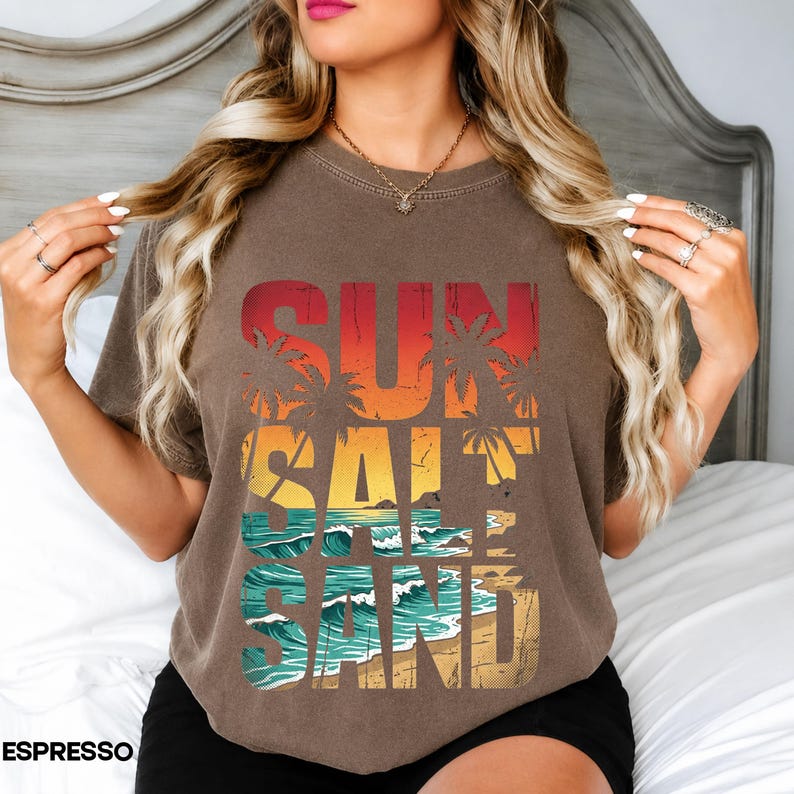 May include: Espresso-colored t-shirt with a beach-themed graphic. The design features the words "SUN SALT SAND" in a layered, colorful design with palm trees, ocean waves, and a sandy beach.