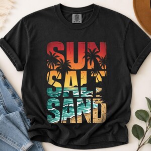 May include: Black t-shirt with a colorful graphic design. The design features the words "SUN SALT SAND" stacked vertically, with a beach scene incorporated into the letters. The colors include red, orange, yellow, teal, and tan.
