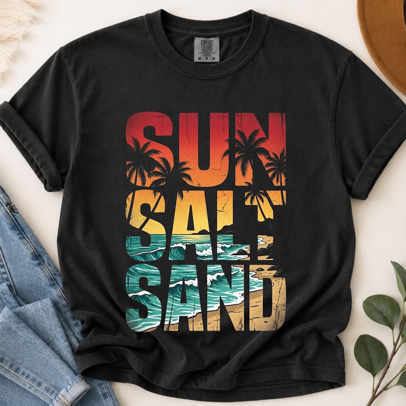 May include: Black t-shirt with a colorful graphic design. The design features the words "SUN SALT SAND" stacked vertically, with a beach scene incorporated into the letters. The colors include red, orange, yellow, teal, and tan.