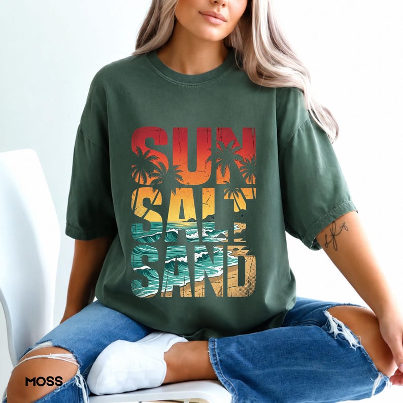 May include: A moss green t-shirt with the words "SUN SALT SAND" in a colorful, beach-themed design. The graphic features palm trees, a sunset, and ocean waves. The shirt is a relaxed fit and is worn by a person.