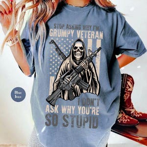 Op de afbeelding: Een blauw spijkerkleurig t-shirt met een afbeelding van een skelet in een kapmantel die een geweer vasthoudt. Achter het skelet staat een vervaagde Amerikaanse vlag. De tekst luidt: STOP ASKING WHY I'M GRUMPY VETERAN I DON'T ASK WHY YOU'RE SO STUPID.