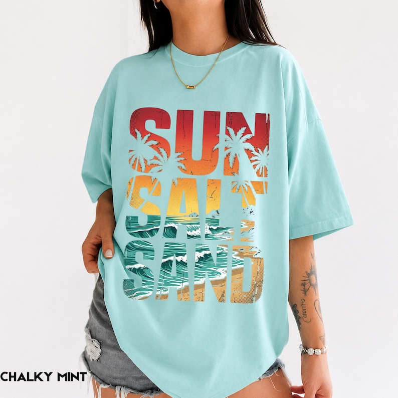 May include: A light blue t-shirt with the words "SUN SALT SAND" in a gradient design of red, orange, yellow, and brown. The design includes palm trees, ocean waves, and a beach scene.