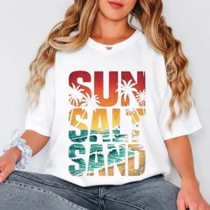 May include: White t-shirt with a colorful graphic design. The design features the words "SUN SALT SAND" in a layered style, with palm trees and a beach scene. The shirt is a casual, relaxed fit.