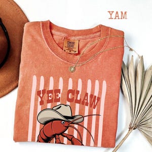 Könnte beinhalten: Orangefarbenes Comfort Colors T-Shirt mit einer Grafik einer Languste mit Cowboyhut und den Worten "Yee Claw". Das Shirt hat vertikale rosa Streifen und eine goldene Halskette. Das Wort "YAM" ist oben aufgedruckt.