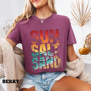 May include: Berry-colored t-shirt with the words "SUN SALT SAND" in a layered design. The design features a sunset, palm trees, ocean waves, and sand. The word "BERRY" is printed at the bottom left.