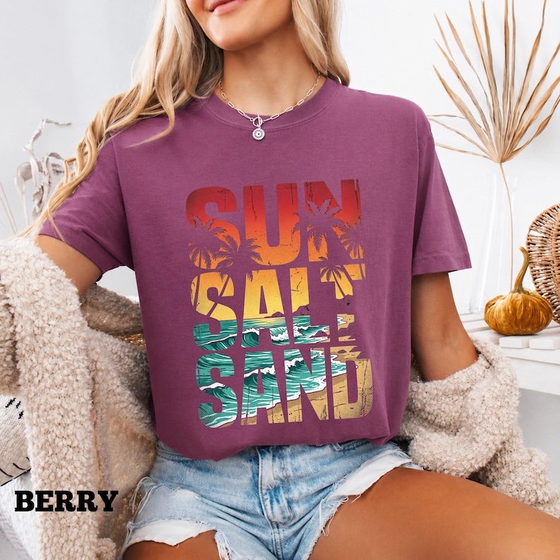 May include: Berry-colored t-shirt with the words "SUN SALT SAND" in a layered design. The design features a sunset, palm trees, ocean waves, and sand. The word "BERRY" is printed at the bottom left.