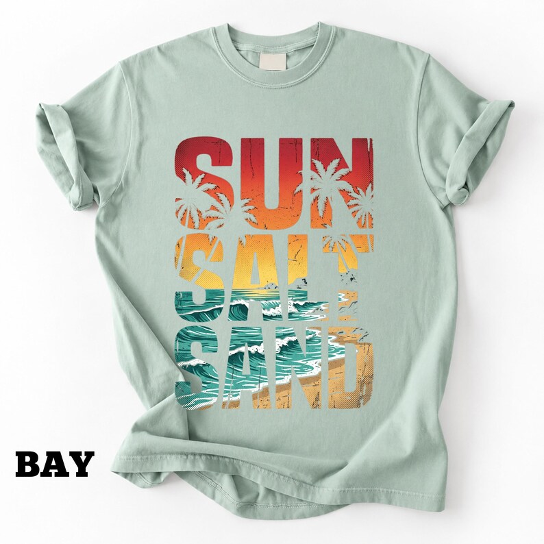 May include: A light green t-shirt with the words "SUN SALT SAND" in a colorful, distressed font. The design includes palm trees, a sunset, and ocean waves, evoking a beach theme.