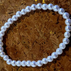 Puede incluir: Un brazalete de perlas blancas. El brazalete está hecho de pequeñas perlas blancas redondas ensartadas juntas. Las perlas tienen un acabado brillante y están dispuestas en un patrón circular. El fondo es una superficie marrón texturizada.