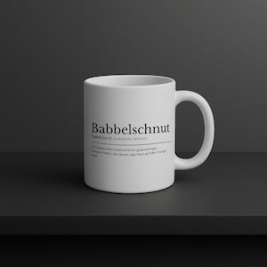 Può includere: Una tazza di ceramica bianca con la parola "Babbelschnut" stampata in nero. La tazza ha anche testo aggiuntivo sotto la parola. La tazza è su una superficie scura.