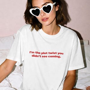 Könnte beinhalten: Weißes T-Shirt mit dem roten Text "I'm the plot twist you didn't see coming." Die Person trägt weiße herzförmige Sonnenbrillen mit schwarzen Gläsern. Der Hintergrund ist eine hellrosa Wand.