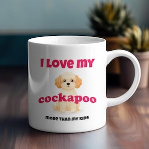 Könnte beinhalten: Weiße Keramik-Tasse mit dem Text "I Love my cockapoo more than my kids" in Rosa und Schwarz. Die Tasse zeigt eine Cartoon-Illustration eines Cockapoo-Hundes. Die Tasse hat einen weißen Henkel und steht auf einer Holzoberfläche.