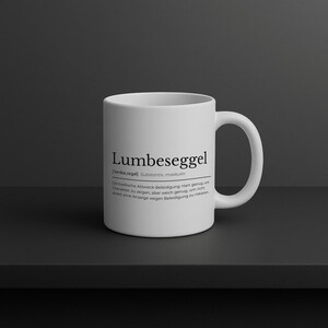 May include: White ceramic mug with the word "Lumbeseggel" printed in black. The mug also features a German definition. The mug is on a dark surface.