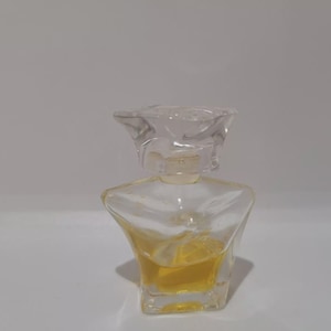 Könnte beinhalten: Eine klare Parfümflasche aus Glas mit einem einzigartigen, eckigen Design. Die Flasche hat einen transparenten, blütenförmigen Verschluss und enthält eine gelbe Flüssigkeit. Die Flasche steht auf einer weißen Oberfläche.