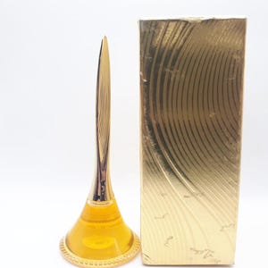 Könnte beinhalten: Eine Vintage-Parfümflasche mit einem goldfarbenen, blattförmigen Verschluss und einer passenden goldenen Schachtel. Die Flasche enthält eine gelbe Flüssigkeit. Die Schachtel hat ein strukturiertes, geschwungenes Design.