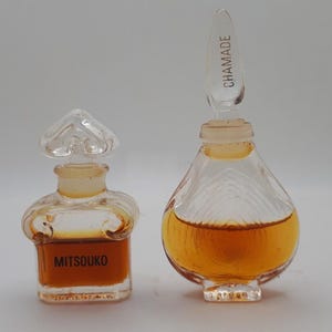 Könnte beinhalten: Zwei Vintage-Parfümflaschen. Die Flasche links ist mit "Mitsouko" beschriftet und hat einen herzförmigen Verschluss. Die Flasche rechts ist mit "Chamade" beschriftet und hat einen tropfenförmigen Verschluss. Beide Flaschen enthalten eine goldfarbene Flüssigkeit.