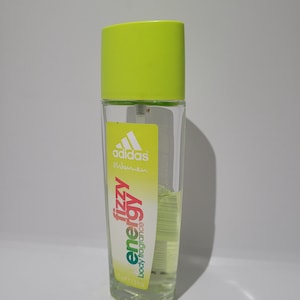 Könnte beinhalten: Eine klare Glasflasche Adidas Fizzy Energy Körperspray. Die Flasche hat einen leuchtend grünen Deckel und ein gelb-grünes Etikett mit dem Adidas-Logo und Produktinformationen. Enthält ca. 75 ml hellgrüne Flüssigkeit.