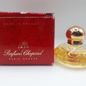 Könnte beinhalten: Ein Vintage-Parfümflakon mit goldfarbenem Verschluss und einem transparenten, gemusterten Glaskörper, gefüllt mit einer gelben Flüssigkeit. Die Flasche steht neben einer roten Schachtel mit dem Text "Parfums Chopard" und "Made in France".