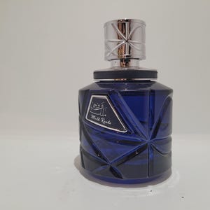 Può includere: Un flacone di profumo blu scuro con un design geometrico. Il flacone ha un tappo argentato e un'etichetta con caratteri arabi e le parole "Milk Roads". Il flacone è su una superficie bianca.