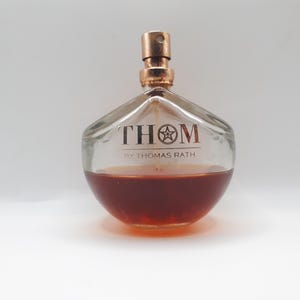Pode incluir: Um frasco de perfume de vidro transparente com um borrifador de cor cobre e um líquido âmbar escuro no interior. O frasco tem um formato único e apresenta a palavra "THOM" com um design de estrela e "BY THOMAS RATH".