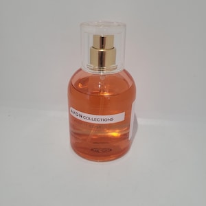 Könnte beinhalten: Eine transparente Parfümflasche mit goldfarbener Pumpe und transparentem Kunststoffverschluss. Die Flasche enthält eine orangefarbene Flüssigkeit und trägt ein weißes Etikett mit dem Text "AVON COLLECTIONS KEEP IT COSY".