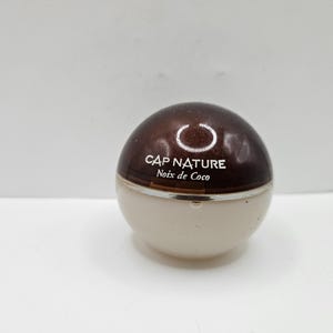 Könnte beinhalten: Ein kleiner, runder Kosmetikbehälter mit zweifarbigem Design. Die Oberseite ist glänzend dunkelbraun, während die Unterseite hellbeige ist. Der Text "CAP NATURE Noix de Coco" ist auf dem Behälter aufgedruckt.