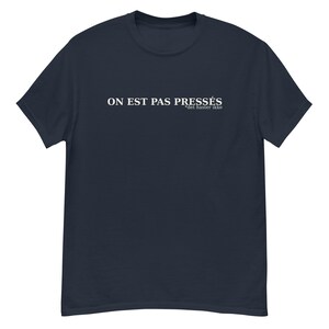May include: Navy blue t-shirt featuring the white text "ON EST PAS PRESSÉS" and "det haster ikke". The shirt has a crew neck and is made of a soft fabric. A casual and comfortable apparel item.