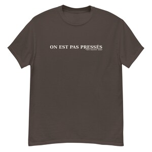 May include: A brown t-shirt featuring the white text "ON EST PAS PRESSÉS" and "det haster ikke". The shirt has a crew neck and short sleeves.