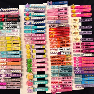 Può includere: Una collezione di mollette decorative in legno con varie frasi e abbellimenti. Le mollette presentano glitter, strass e ciondoli, con testi come "BEAST MODE", "SPARKLE" e "ROCKSTAR". I colori includono rosa, blu, giallo e nero.
