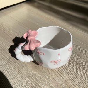 Puede incluir: Una taza de cerámica blanca con un asa negra y el texto "But first, coffee" en un estilo manuscrito. La taza está sobre una superficie blanca. La taza mide 10 cm de alto.