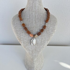 May include: A necklace with brown, rectangular beads and silver accents. The necklace features a rectangular pendant with a face design. The necklace is displayed on a mannequin.