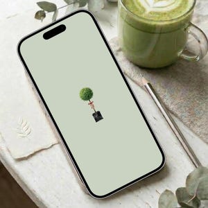 May include: A smartphone displays a light green screen with a small, centered illustration of a topiary tree in a black pot, tied with a red ribbon. A glass mug of green beverage and a pencil are visible in the background.