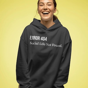 May include: Black hooded sweatshirt with the text "ERROR 404 Social Life Not Found" in white. The sweatshirt has a drawstring hood and a front pocket. The background is a solid yellow color.