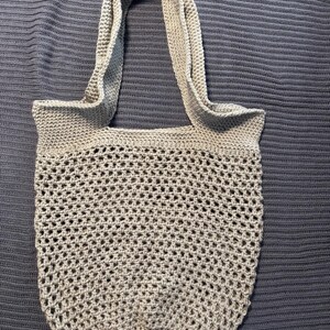 May include: A light gray crocheted tote bag with a wide, open weave design. The bag has a rounded bottom and two long handles. The bag is made of a soft, textured yarn.