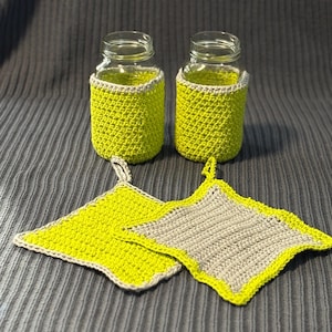 May include: Two glass jars with bright green crochet cozies and two matching square pot holders. The pot holders are two-toned, with a green center and a gray border, and a loop for hanging. The cozies and pot holders are handmade.