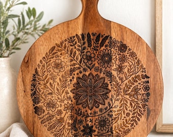 Puede incluir: Tabla de cortar redonda de madera con asa, con un diseño floral detallado grabado en la madera. La tabla es de color marrón cálido, con un patrón floral circular en el centro. El asa tiene un agujero para colgar.