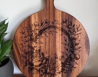 Puede incluir: Tabla de cortar redonda de madera con asa y diseño de corona floral. La tabla es de madera marrón claro y presenta un patrón floral grabado con láser. El asa tiene una abertura circular. El diseño floral incluye peonías y otros elementos botánicos.