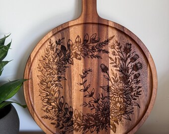 Puede incluir: Tabla de servir redonda de madera con asa. La tabla presenta un diseño de corona floral grabado en la madera. La veta de la madera es visible y la tabla tiene un color marrón cálido natural. El asa tiene un agujero para colgar.