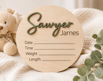 May include: A circular light-colored wooden birth announcement sign featuring the name Sawyer James in raised dark green script and block lettering. Below the name are printed lines for Date, Time, Weight, and Length to record baby details.