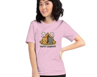 May include: A light pink t-shirt featuring a cartoon illustration of two cats inside a tent with the text "HAPPY CAMPERS" below. The cats are a light orange and gray color. The tent is brown with green grass and white clouds.