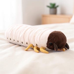 May include: A plush caterpillar toy in cream and brown. The toy has a segmented body with dark brown spots and a rounded brown head. It features small yellow legs and is lying on a white surface.