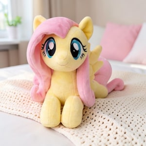 May include: A plush toy pony with a yellow body, pink mane and tail, and large blue eyes. The toy is sitting on a cream-colored blanket. The pony has a friendly expression and is a popular children's toy.