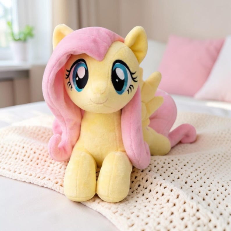May include: A plush toy pony with a yellow body, pink mane and tail, and large blue eyes. The toy is sitting on a cream-colored blanket. The pony has a friendly expression and is a popular children's toy.