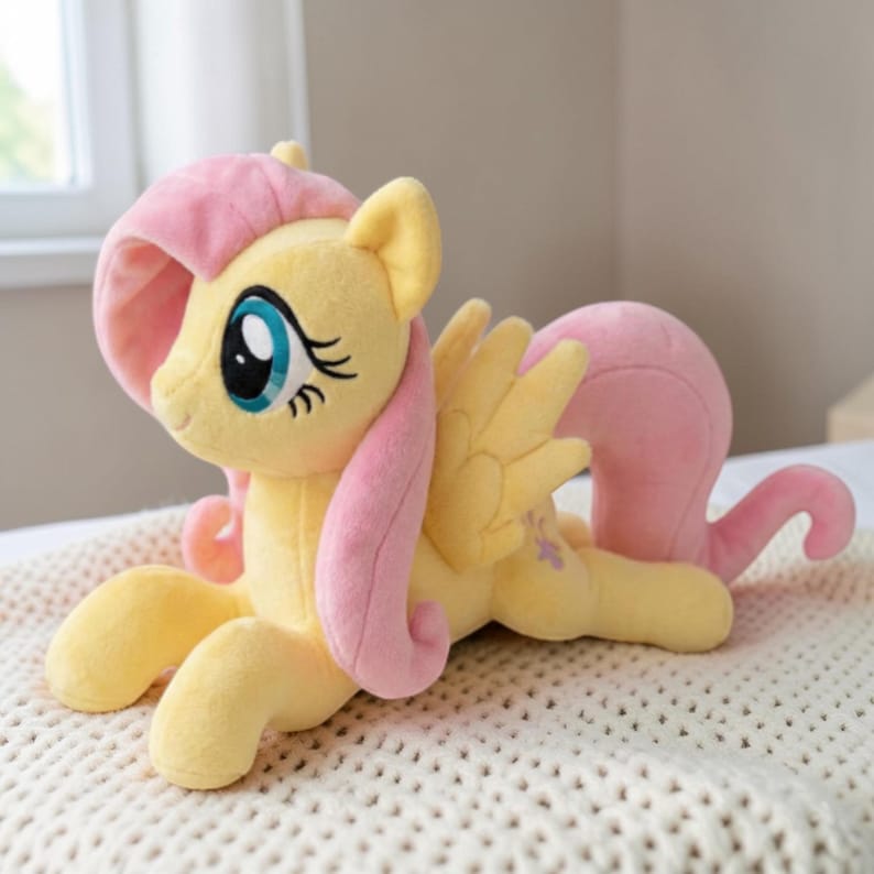 May include: A plush yellow pony with pink hair and wings, lying on a cream-colored blanket. The pony has large blue eyes with black eyelashes and a pink tail. The wings are yellow with purple accents.