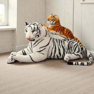 Op de afbeelding: Twee pluche tijgerspeelgoed. Een grote witte tijger met zwarte strepen ligt, en een kleinere oranje tijger met zwarte strepen ligt erop. Het speelgoed staat in een kamer met een raam en lichtgekleurde muren.
