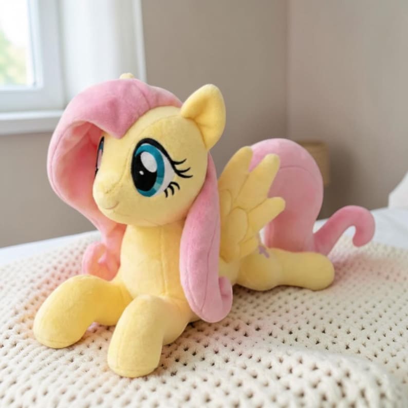 May include: A plush toy pony in a lying position, featuring a yellow body, pink mane and tail, and large blue eyes. The toy has yellow wings and is resting on a cream-colored blanket. The pony is a popular character from a children's animated series.